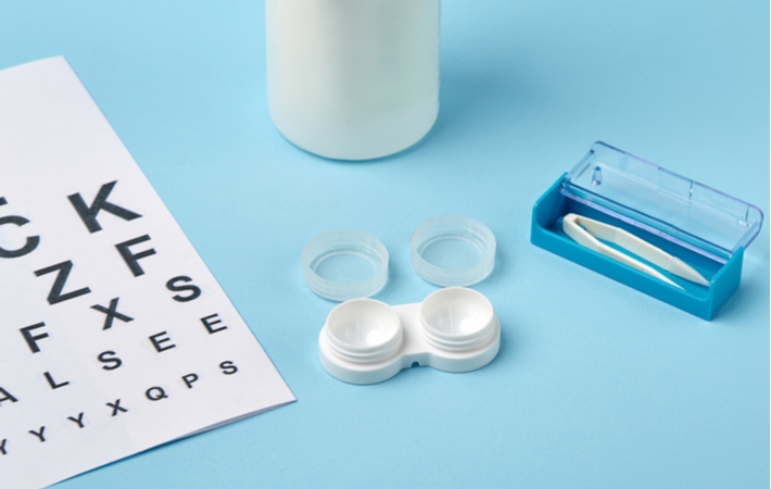 A container of contact lenses placed next to a contact lens applicator and Snellen vision test chart all on a light blue surface