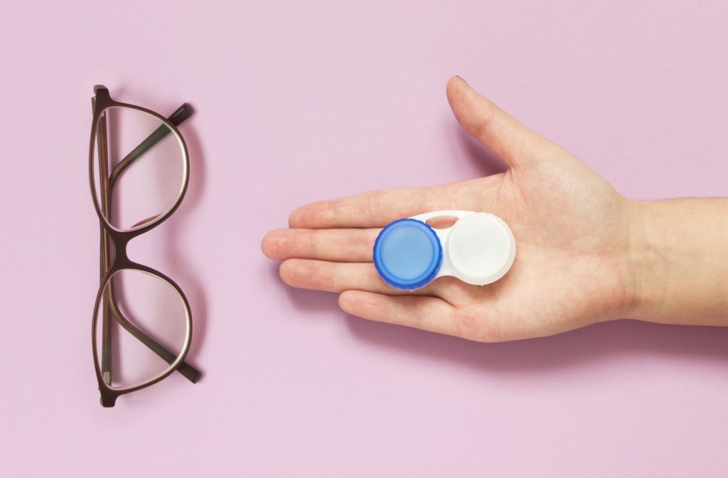 A pair of glasses laying next to someone's arm reaching outright over a pink surface with a set of contact lenses in the storage container resting in their palm