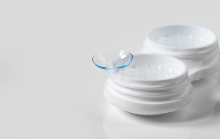 A single contact lens resting on the edge of its container that's sitting on a white surface