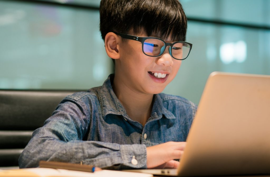 A young boy smiling, wearing his blue light glasses while he uses his laptop at a desk