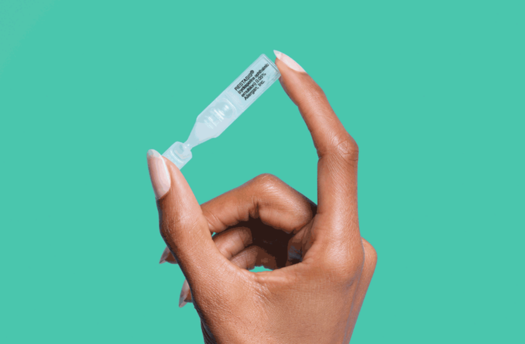 A woman holding onto a dose of Restasis eye drops against a sea green background.