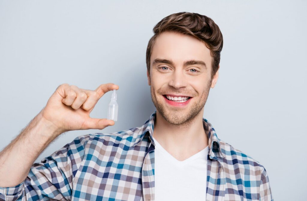 Young man against a grey background holding up a bottle of eye drops between his two fingers and smiling