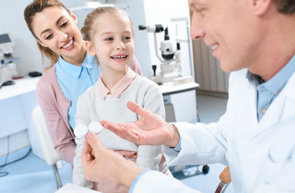 A woman sitting with her young daughter at the optometrist's office. A male optometrist is holding onto a pair of contact lenses while talking to the mother and daughter.