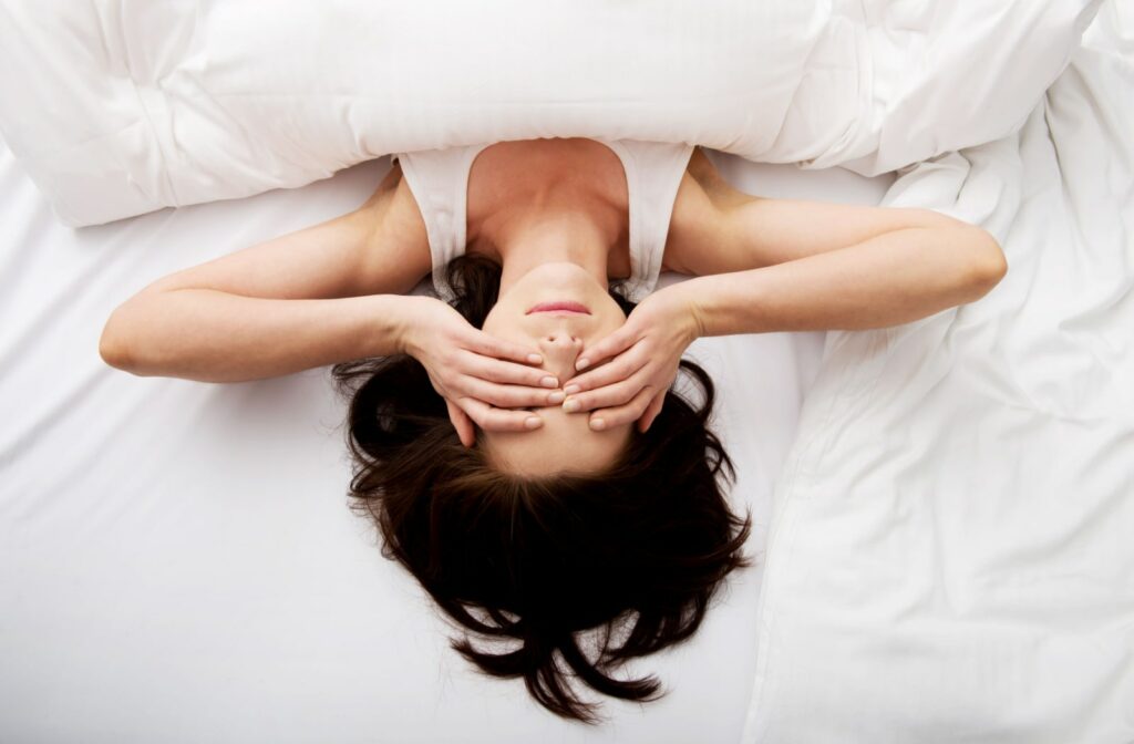 A woman just waking up and lying in bed and she has both of her hands over her eyes